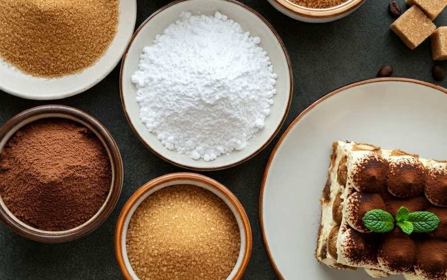 Bols de différents sucres et de cacao vus de dessus, morceaux de sucre et grains de café, avec une part de tiramisu