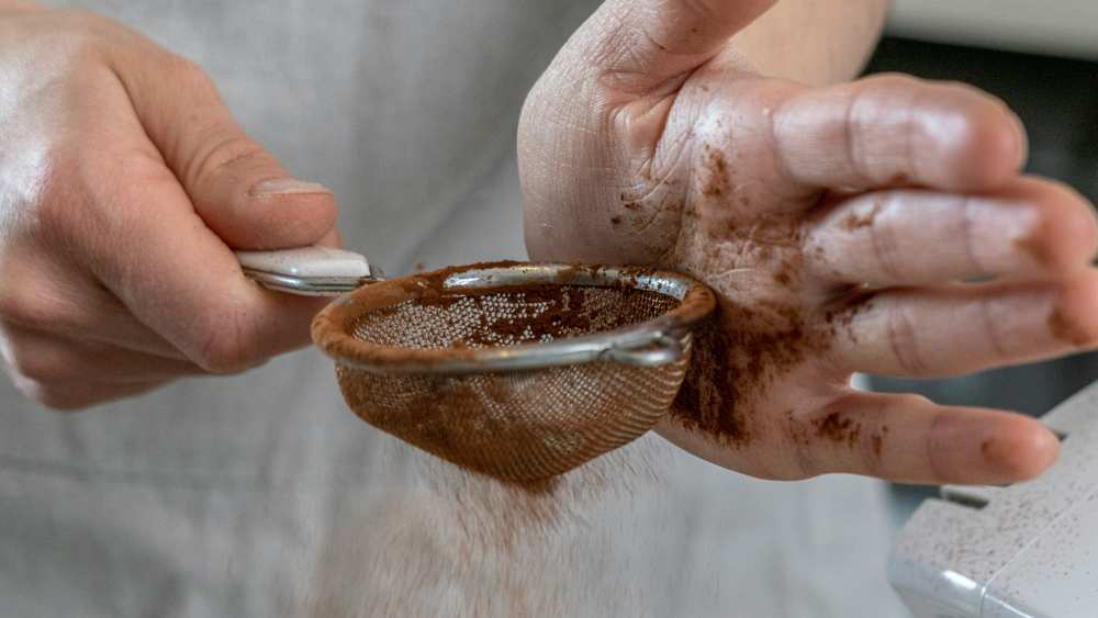 Mains saupoudrant du cacao à travers une petite passoire fine, en tapotant au-dessus d'un saladier