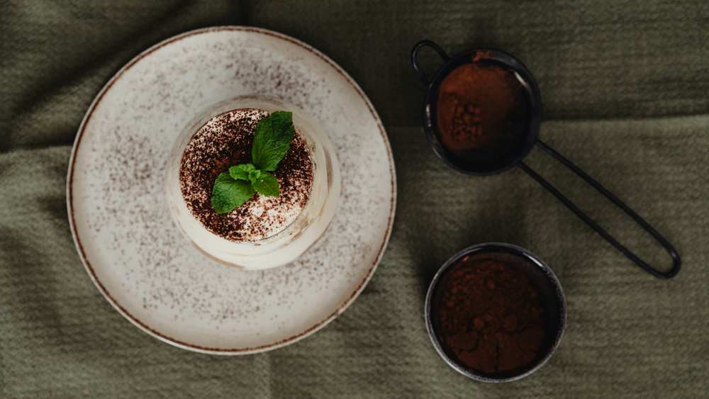 Tiramisu individuel en verre, saupoudré de cacao et décoré de menthe, sur une assiette mouchetée, tamis et bol de cacao à côté