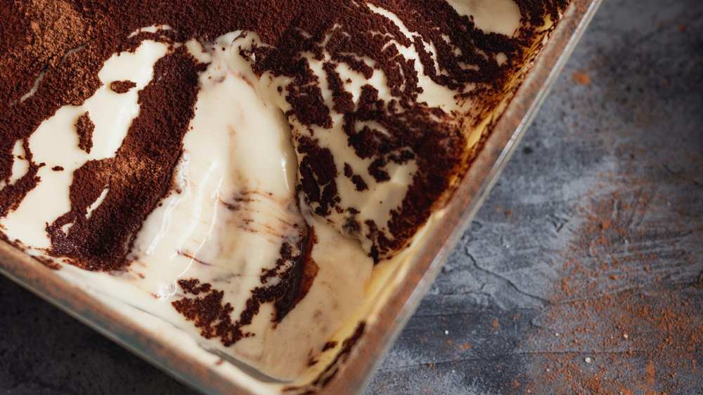 Tiramisu dans un plat en verre saupoudré de cacao, une portion prélevée révélant la crème mascarpone, sur fond gris