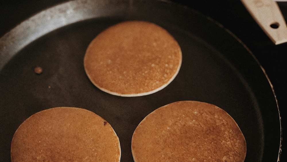 Trois pancakes bien ronds et dorés qui cuisent dans une poêle noire, avec un bol de pâte à côté
