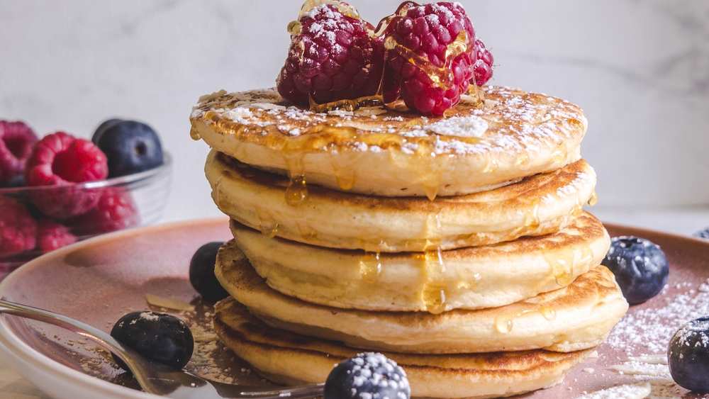 Pile de pancakes moelleux dorés sur une assiette rose, garnis de framboises et de myrtilles, arrosés de sirop