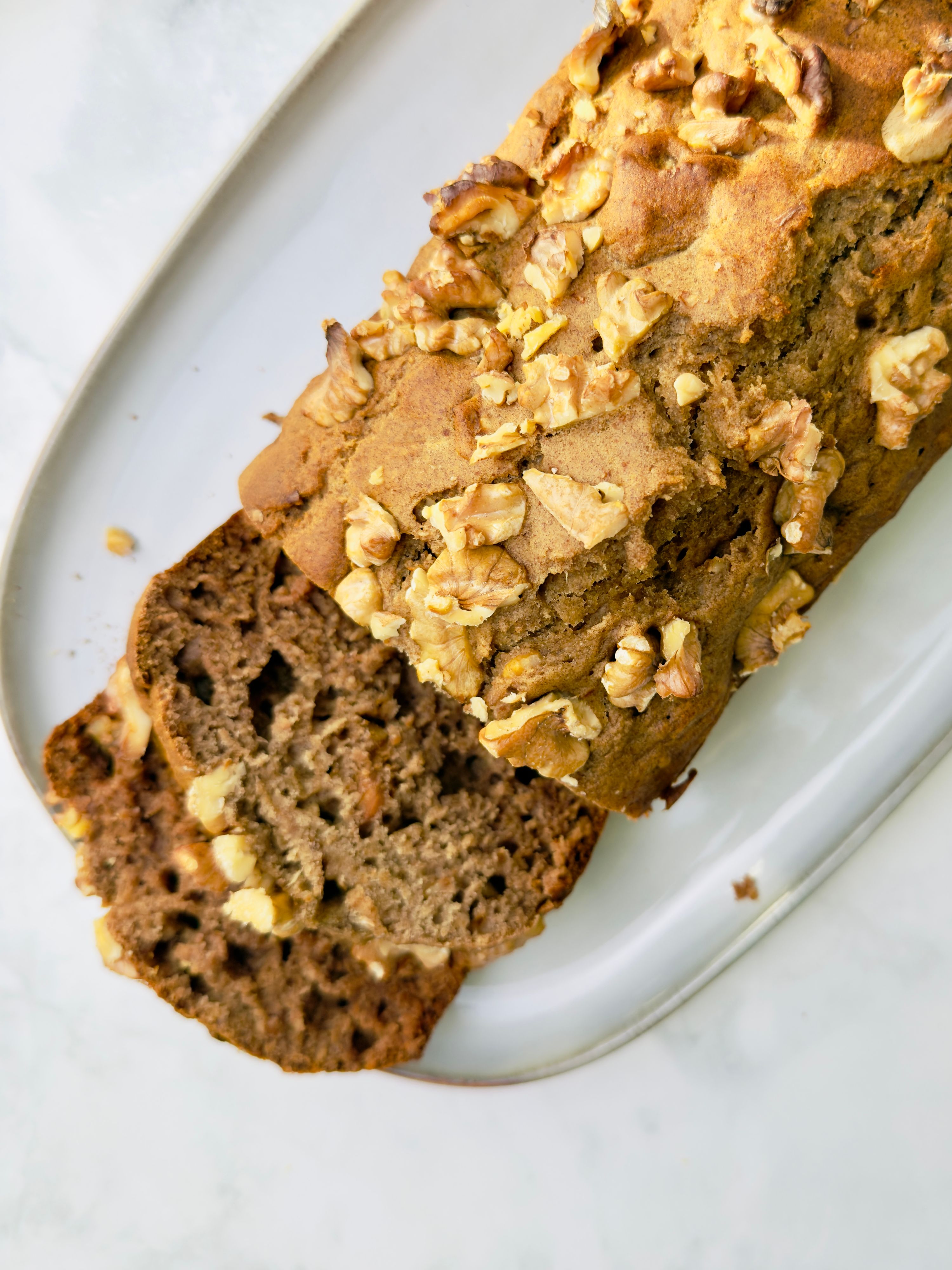 Overhead close-up of a sliced banana bread loaf with walnuts, on a white oval platter over a marble surface.