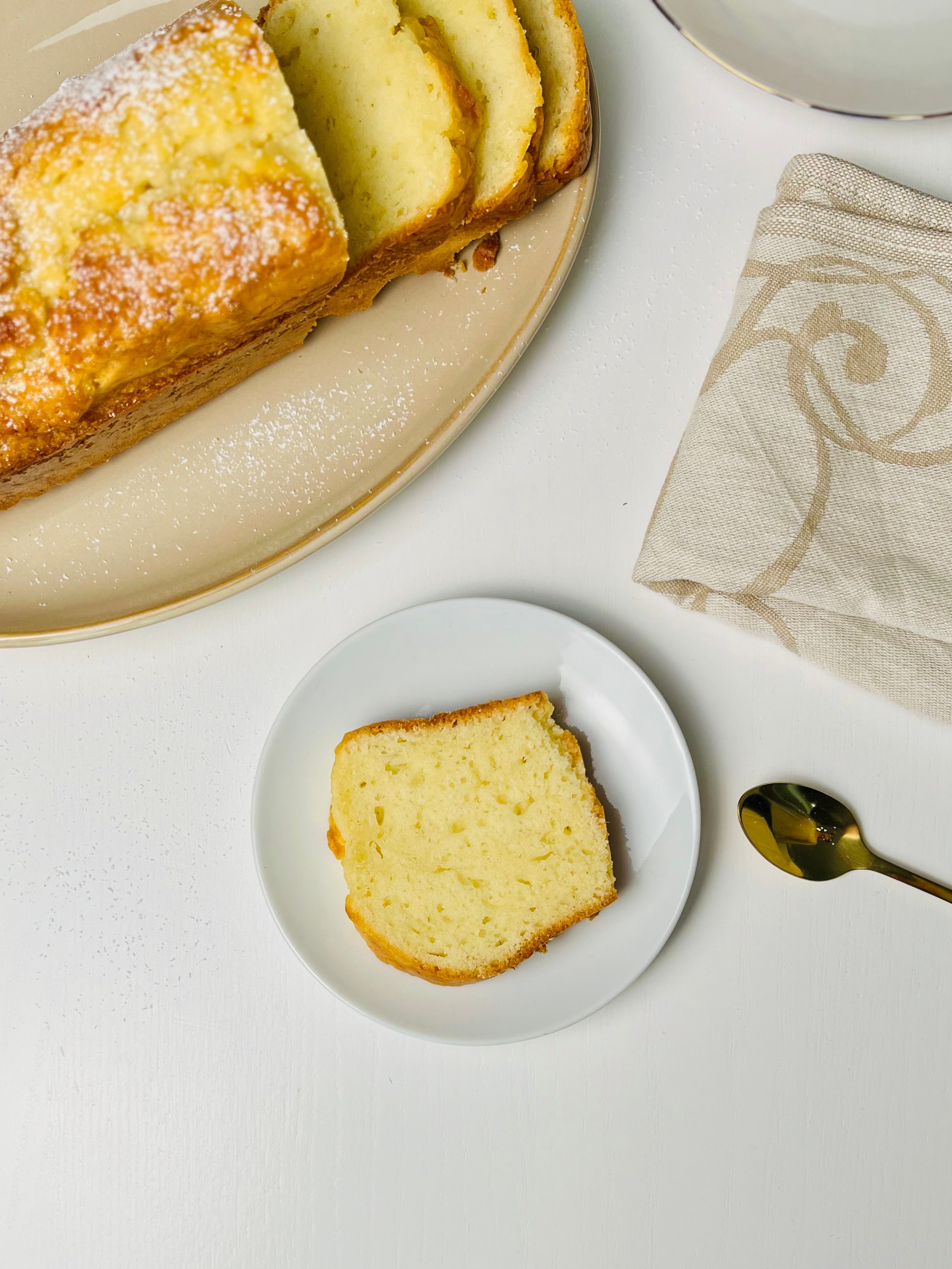 Overhead view of a loaf of cake, partially sliced, and an individual slice, on a white surface with spoon and napkin.