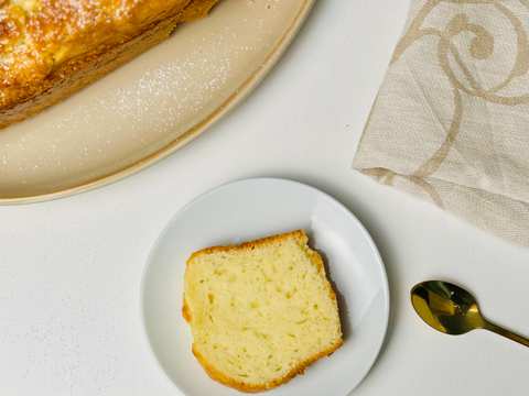 Overhead view of a loaf of cake, partially sliced, and an individual slice, on a white surface with spoon and napkin.