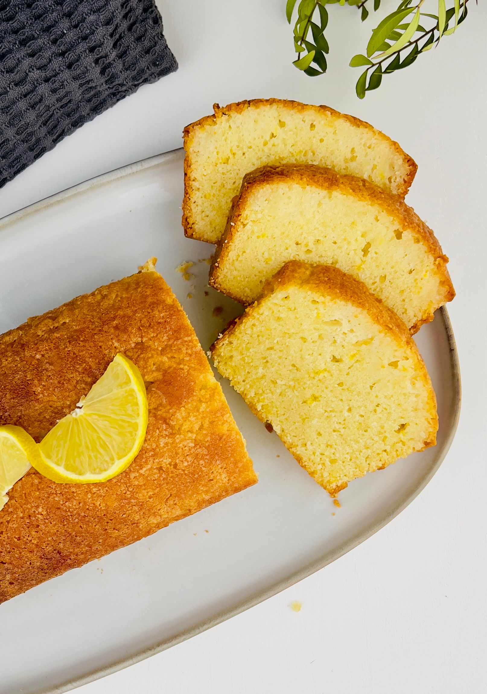 Close-up of a sliced lemon loaf cake on a white rectangular plate. One loaf and three slices of cake, garnish with lemon slices.