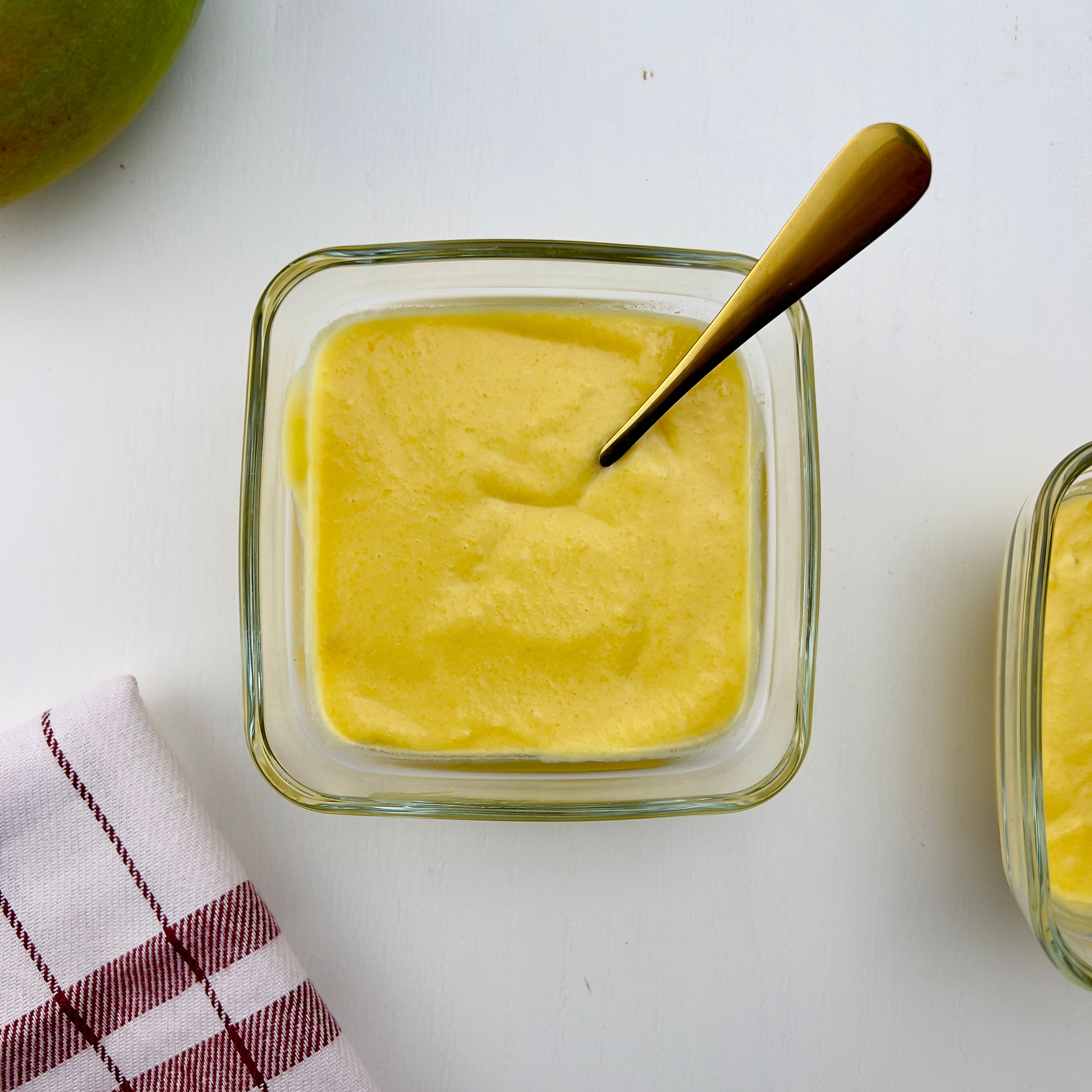 Overhead shot of two square glass bowls with bright yellow mango pudding, one with a gold spoon, and a red and white checkered towel, on a white table.