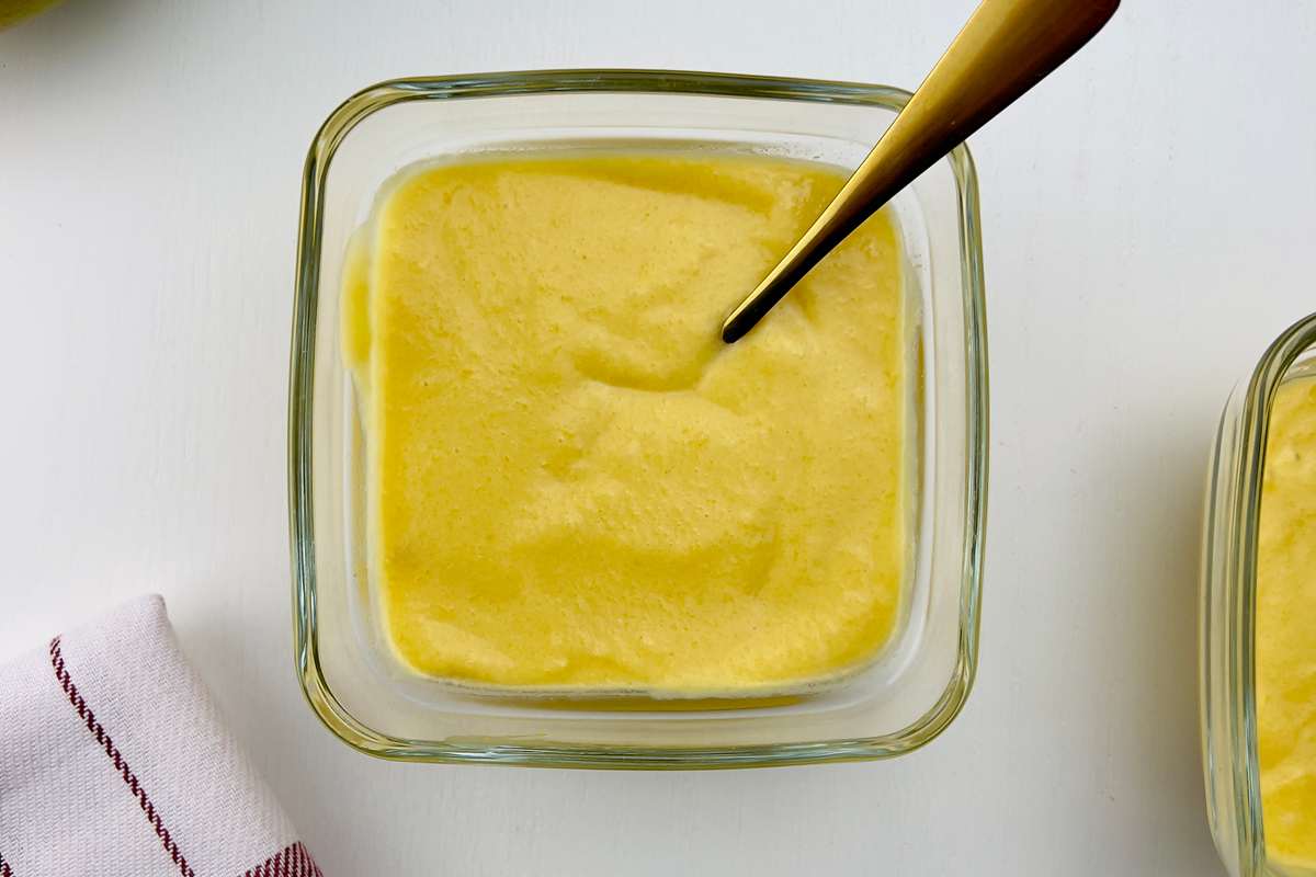 Overhead shot of two square glass bowls with bright yellow mango pudding, one with a gold spoon, and a red and white checkered towel, on a white table.