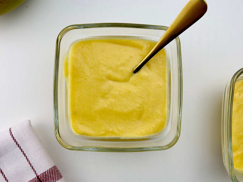 Overhead shot of two square glass bowls with bright yellow mango pudding, one with a gold spoon, and a red and white checkered towel, on a white table.