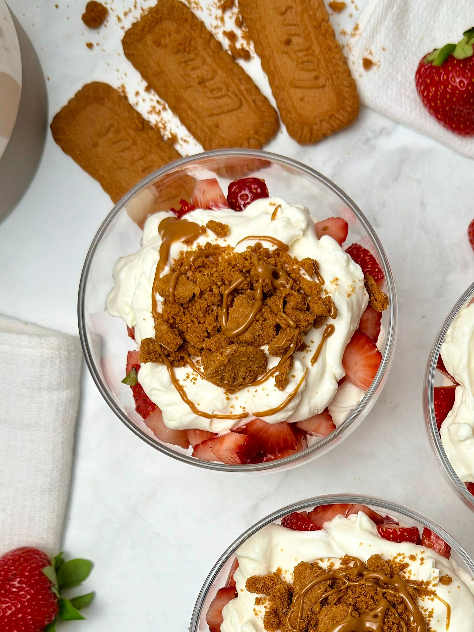 Close up overhead shot of 3 transparent dessert glasses filled with layers of red chopped strawberries, white cream, crushed biscoff cookies, and biscoff sauce. Some biscoff cookies are scattered around the glasses on a white surface.