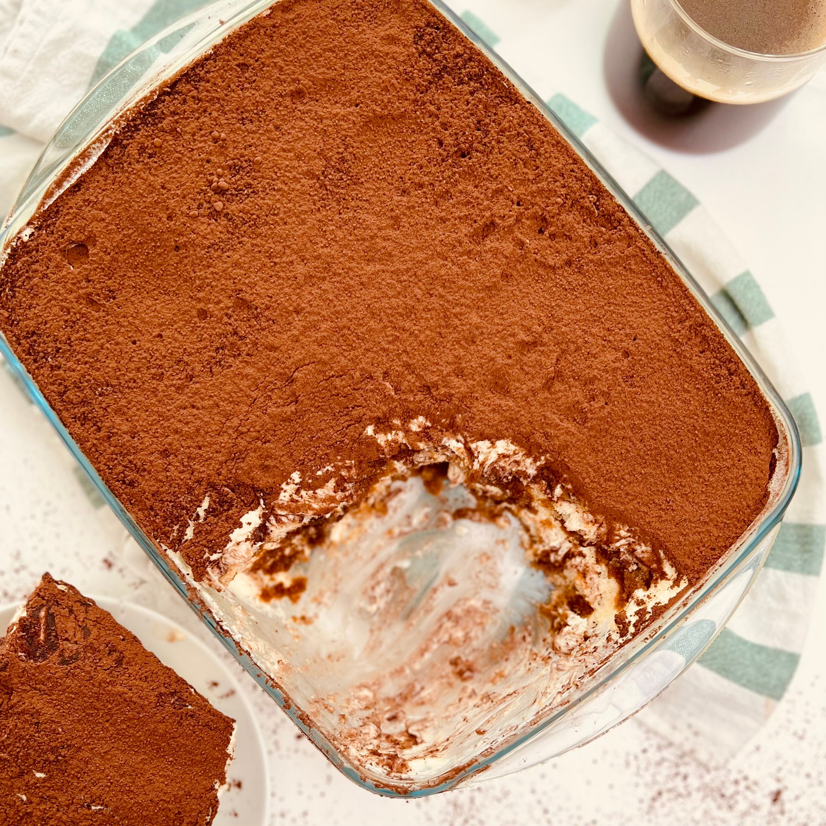 Overhead shot of a glass dish with partly eaten tiramisu, a slice on a plate, and a coffee glass next to it. Messy.