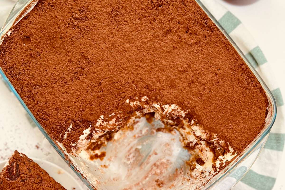 Vue de dessus d'un plat en verre contenant du tiramisu à moitié mangé, une tranche dans une assiette et un verre de café à côté. Désordonné.