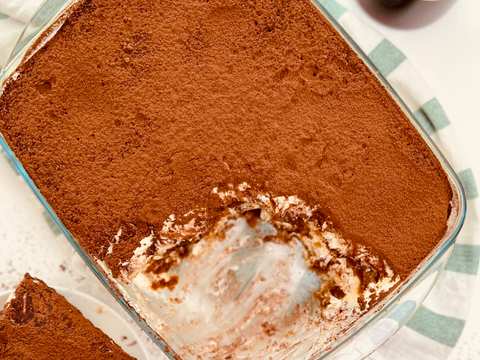Overhead shot of a glass dish with partly eaten tiramisu, a slice on a plate, and a coffee glass next to it. Messy.