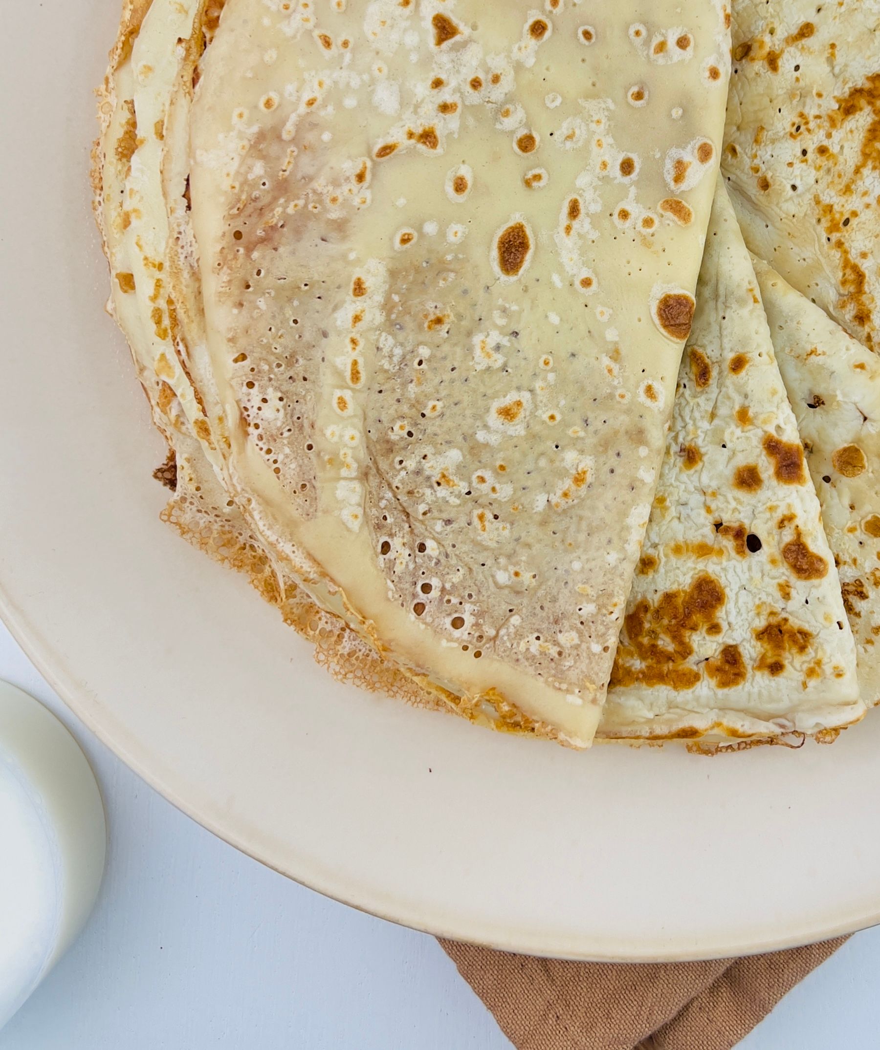 Overhead close-up of a stack of folded pancakes with golden-brown spots on a light plate, with a glass of milk visible in the bottom-left.