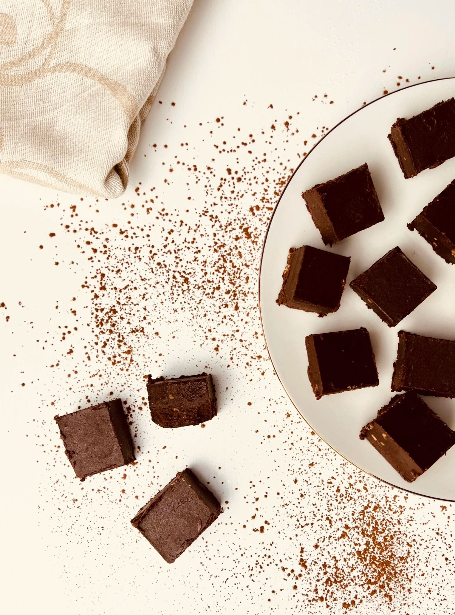 Overhead shot of chocolate fudge squares on a white plate with black and gold rim, scattered cocoa powder, and a napkin.