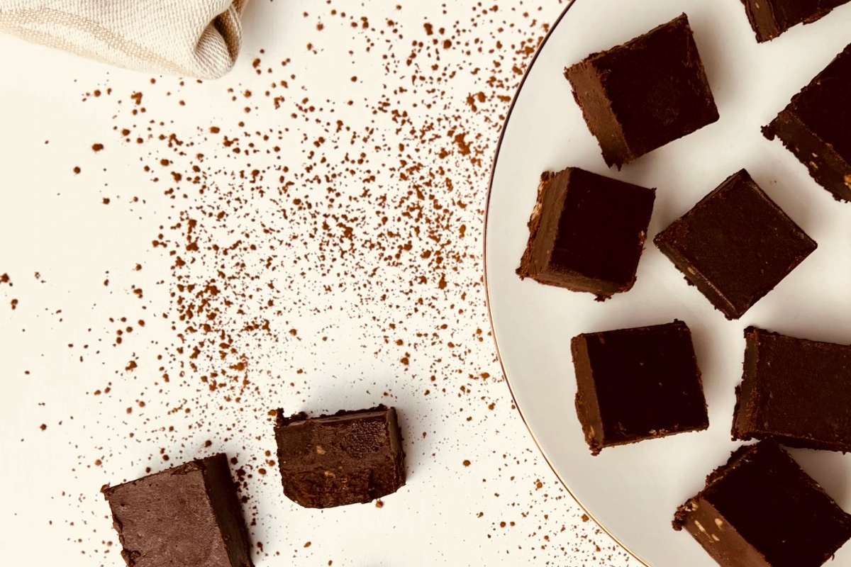 Overhead shot of chocolate fudge squares on a white plate with black and gold rim, scattered cocoa powder, and a napkin.