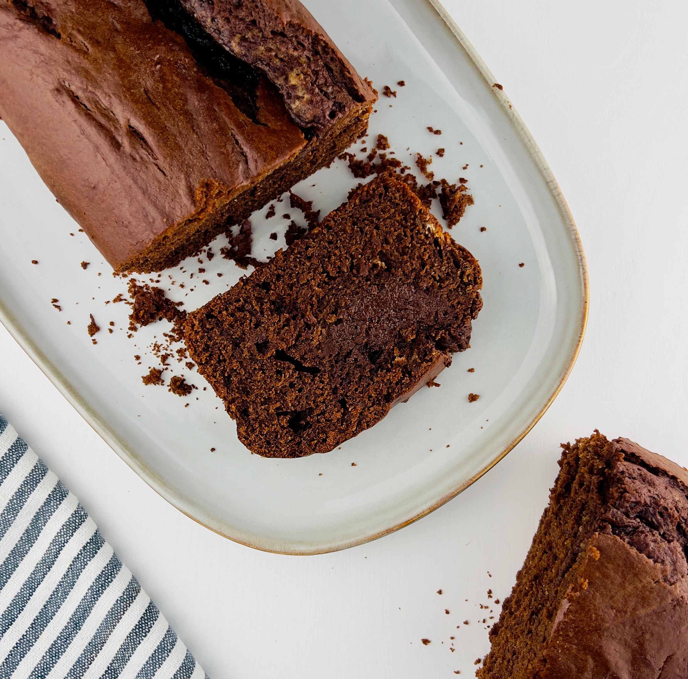 Close-up of a dark chocolate cake loaf partially sliced. A slice sits on a white rectangular ceramic plate beside the loaf with crumbs.