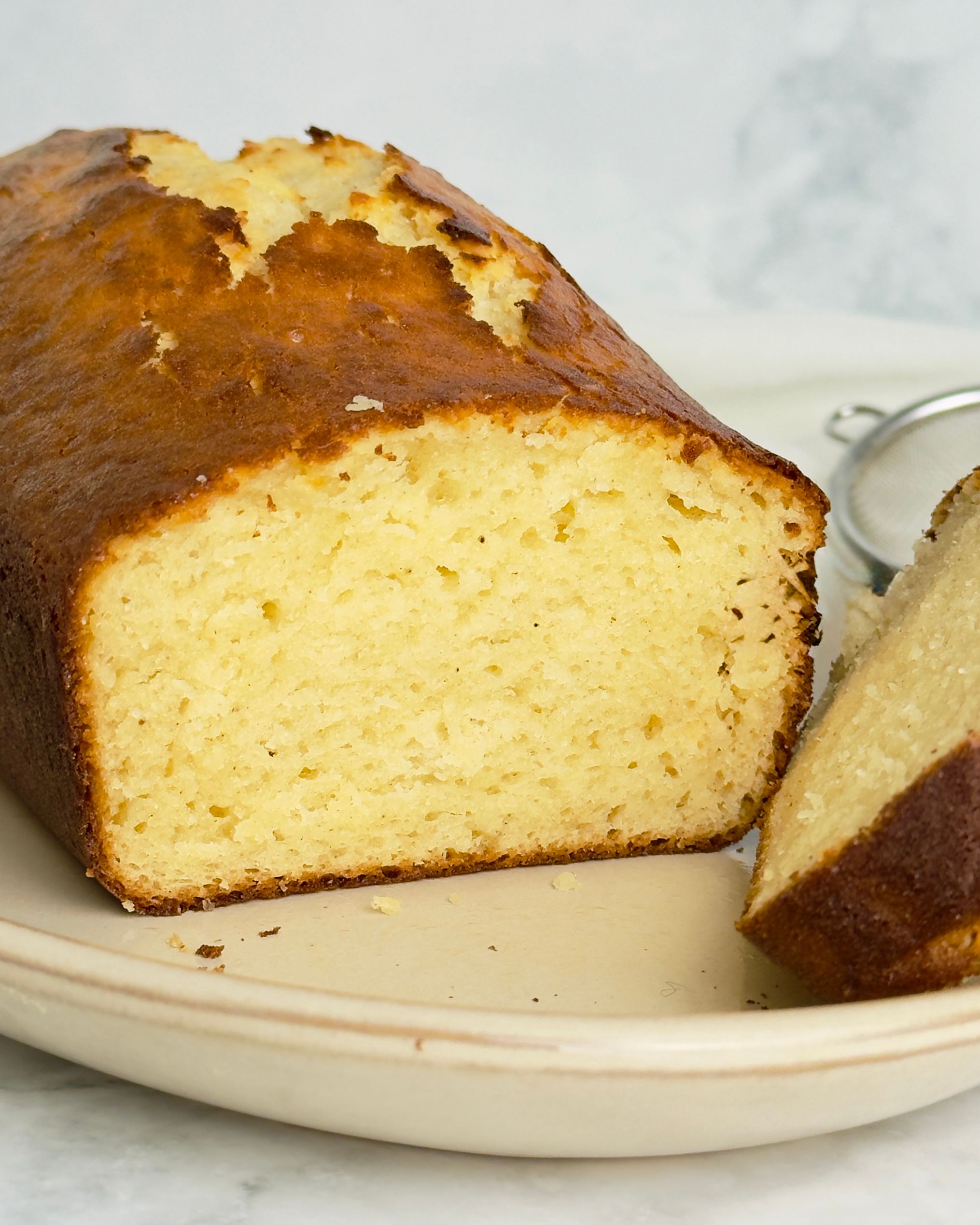 Close-up of a loaf cake with golden crust on a beige plate. Part of the loaf is sliced, showing the light yellow crumb.