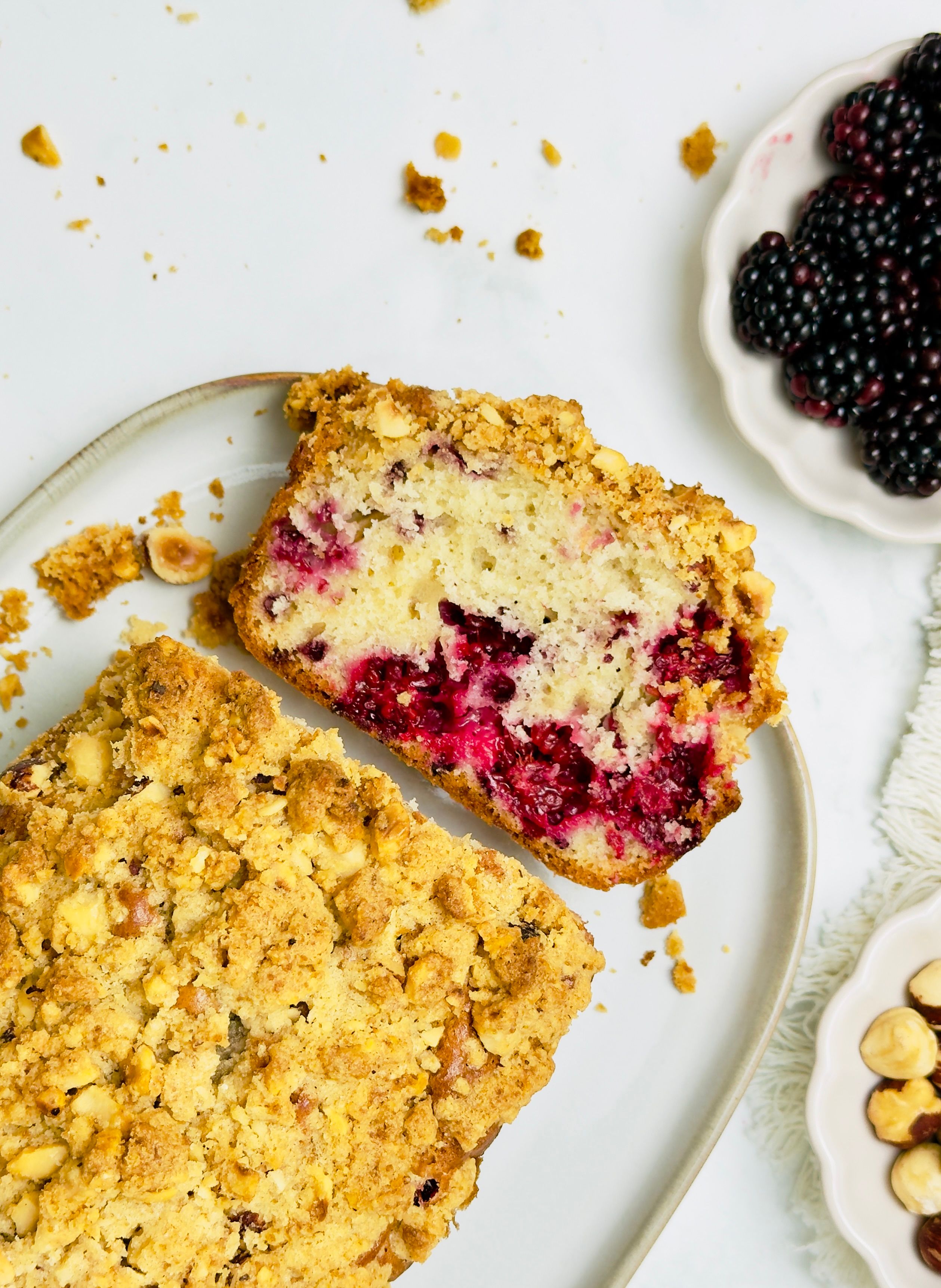 Overhead shot of two pieces of berry and hazelnut streusel cake on a white plate with fresh blackberries and hazelnuts in bowls.