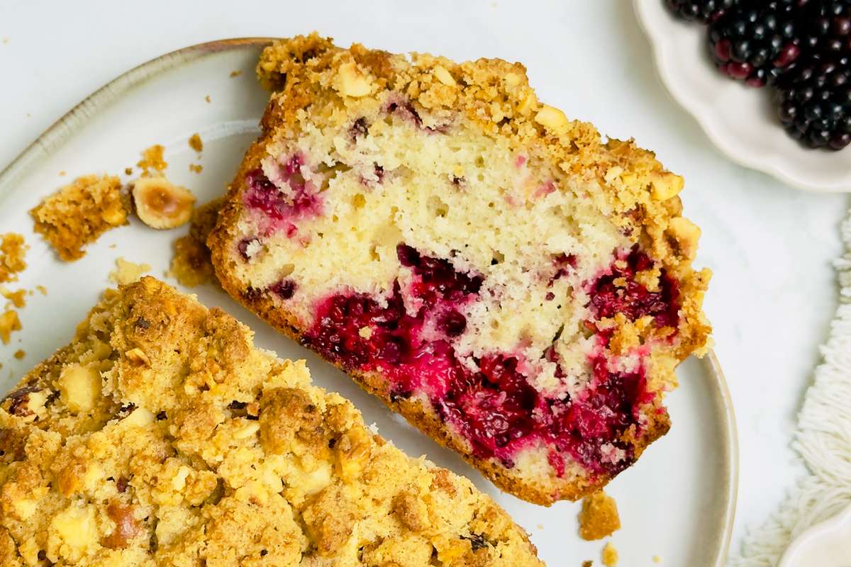 Overhead shot of two pieces of berry and hazelnut streusel cake on a white plate with fresh blackberries and hazelnuts in bowls.