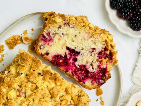 Overhead shot of two pieces of berry and hazelnut streusel cake on a white plate with fresh blackberries and hazelnuts in bowls.