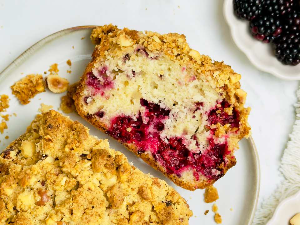 Overhead shot of two pieces of berry and hazelnut streusel cake on a white plate with fresh blackberries and hazelnuts in bowls.