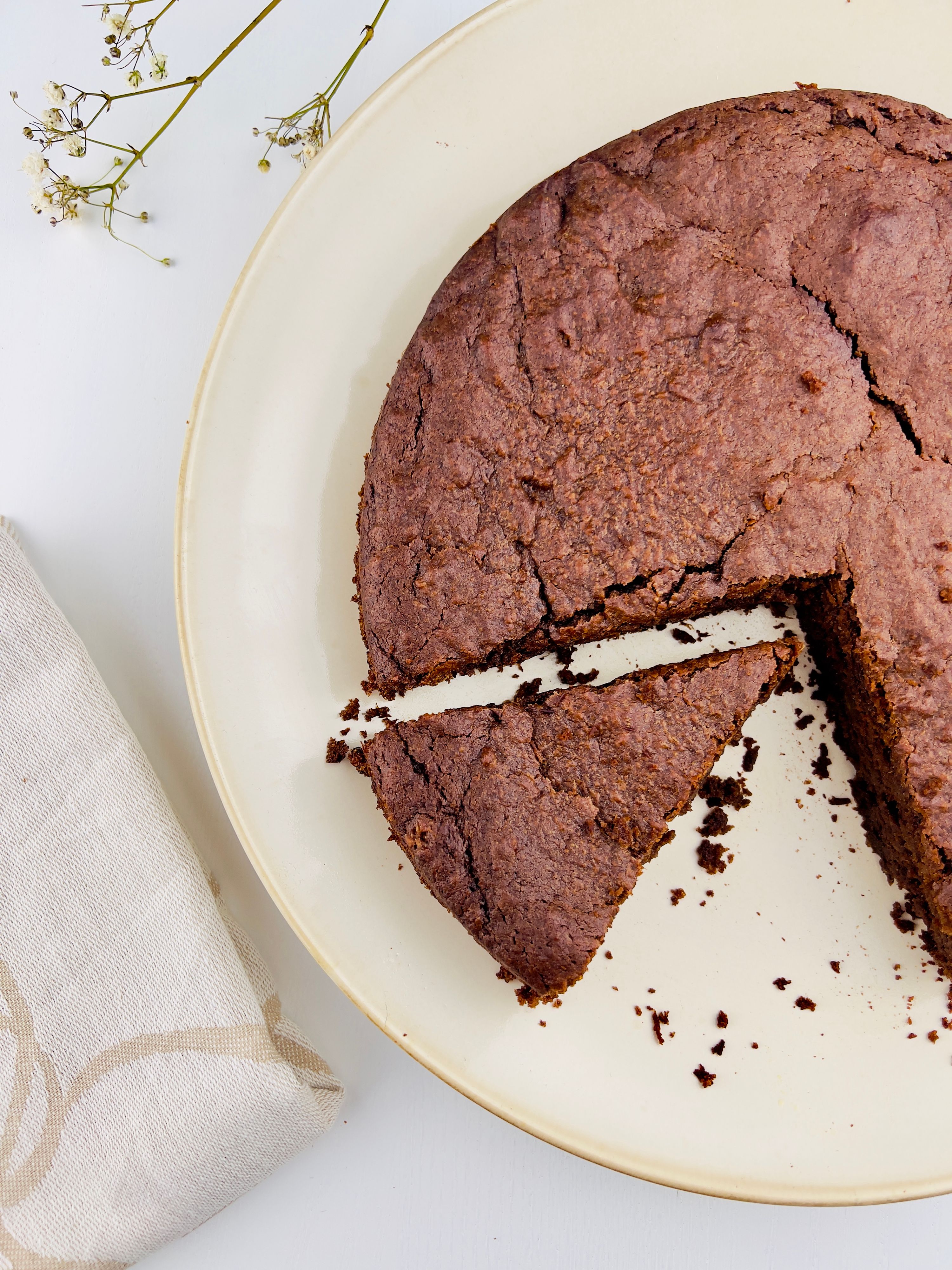 Close-up top-down view of a round chocolate cake on an off-white plate, with one slice removed. Crumbly texture on top. Off-white linen napkin and baby's breath flowers are in the background.