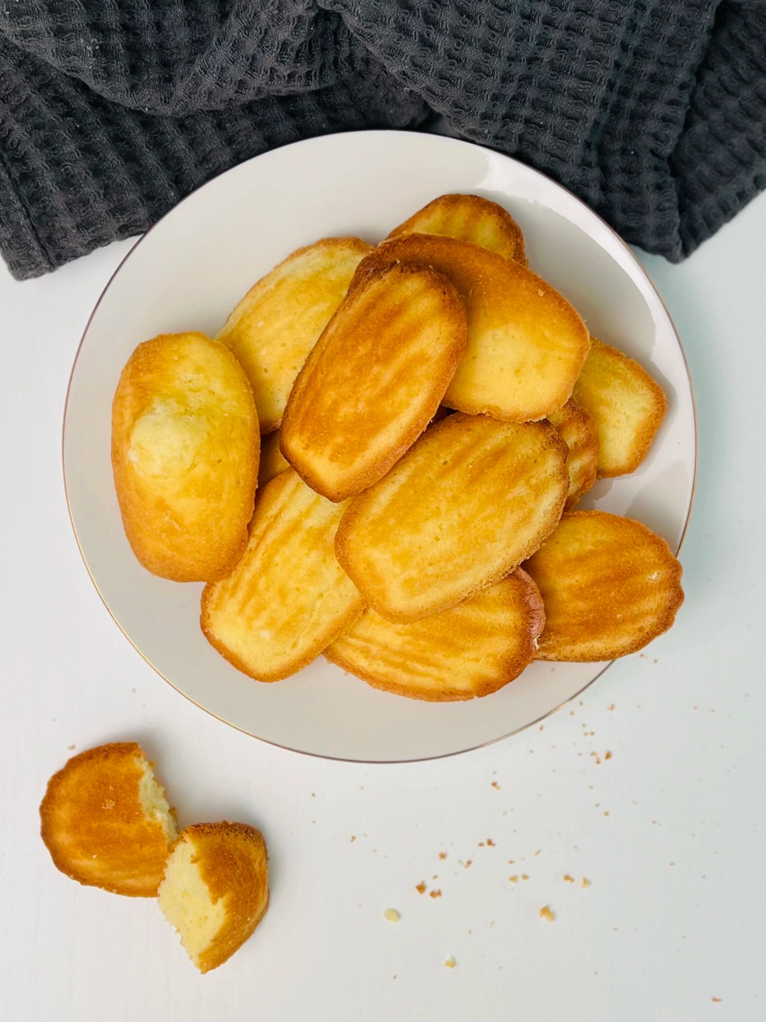 Overhead view of golden-brown shell-shaped madeleines piled on a white plate with a gold rim, one broken madeleine with crumbs beside the plate, dark grey waffle-weave cloth.