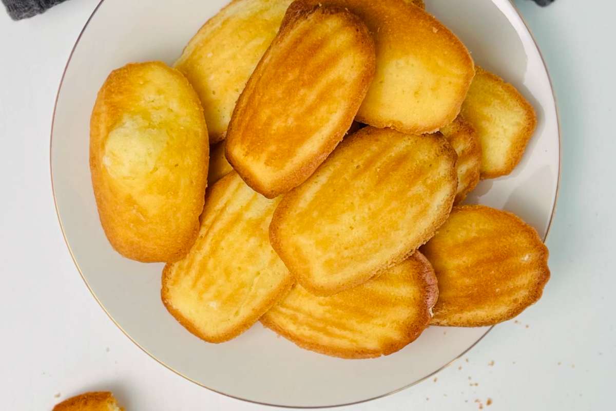 Overhead view of golden-brown shell-shaped madeleines piled on a white plate with a gold rim, one broken madeleine with crumbs beside the plate, dark grey waffle-weave cloth.