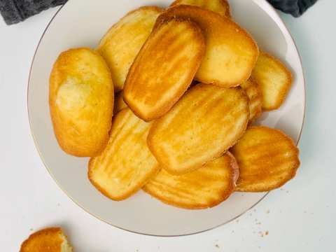 Overhead view of golden-brown shell-shaped madeleines piled on a white plate with a gold rim, one broken madeleine with crumbs beside the plate, dark grey waffle-weave cloth.