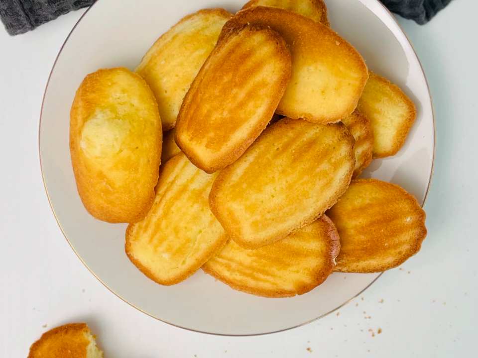 Vue de dessus de madeleines dorées en forme de coquillage empilées sur une assiette blanche à liseré doré, une madeleine cassée avec des miettes à côté, un torchon gris foncé gaufré.