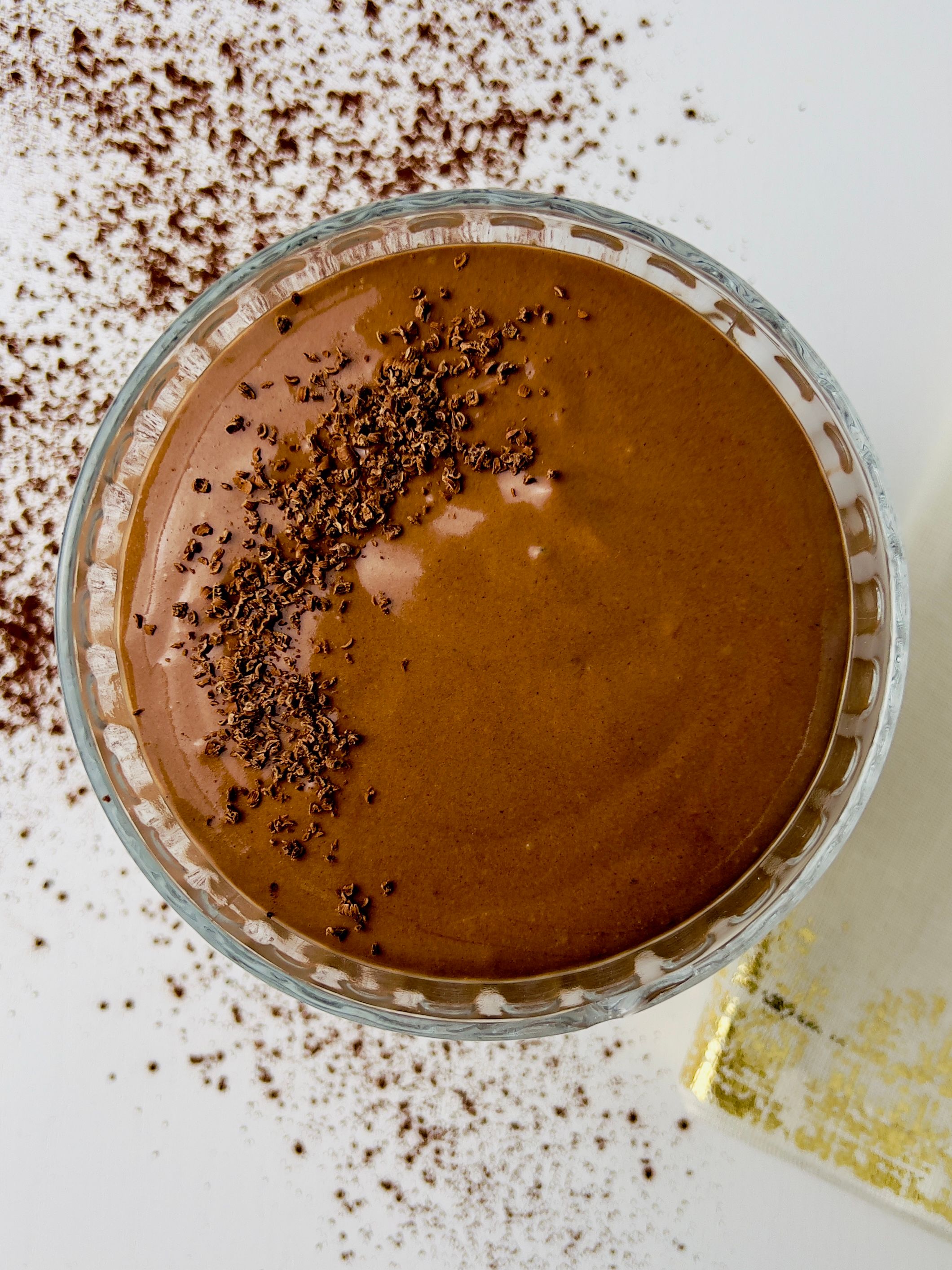 Overhead view of a glass bowl of chocolate mousse topped with chocolate shavings, with cocoa powder sprinkled on the white surface.