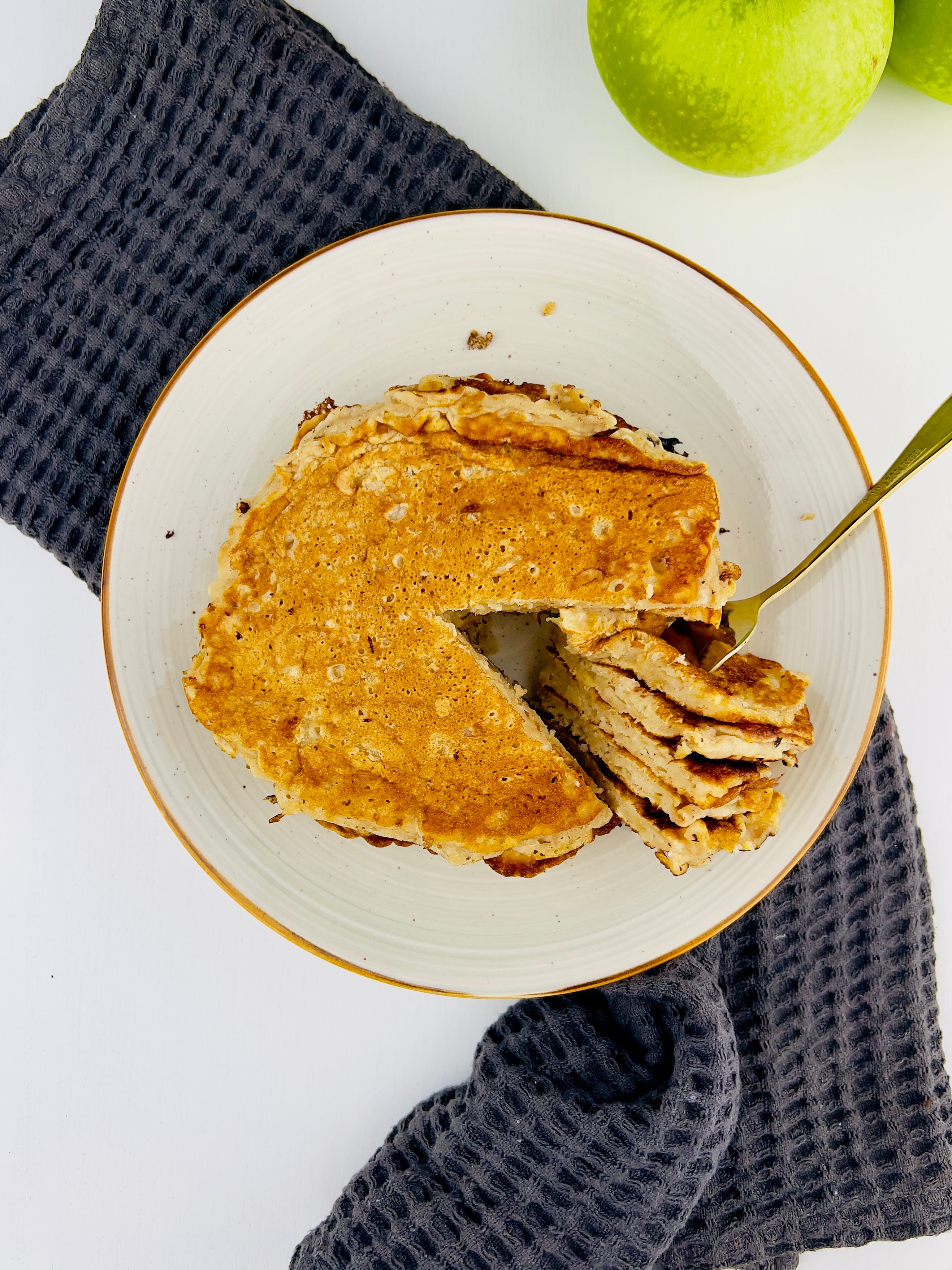 Overhead shot of a plate of apple pancakes on a white surface with a dark grey textured cloth and two green apples.