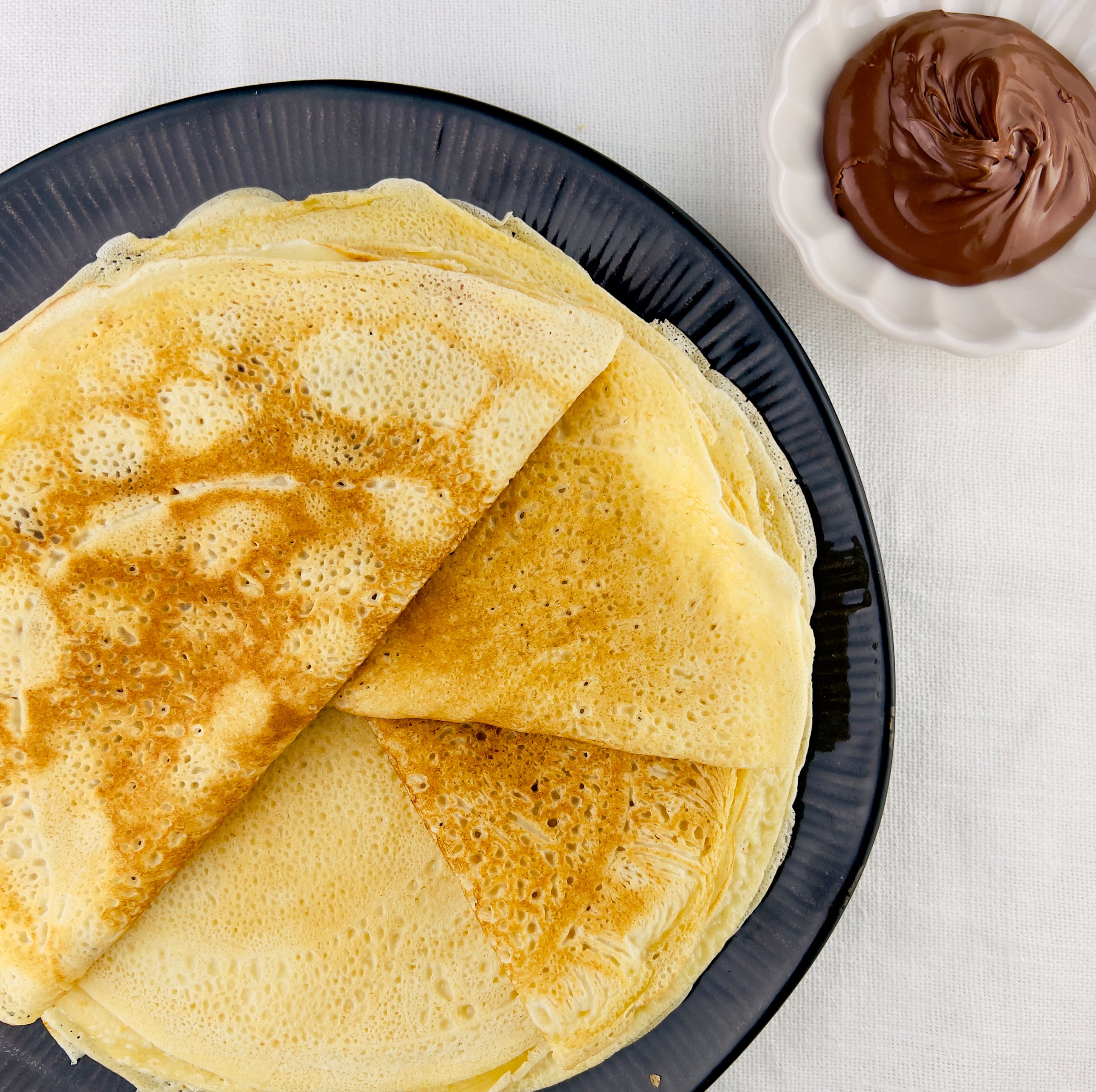 Stacked folded crepes on a black plate with a white scalloped bowl of Nutella to the upper right, all on a white tablecloth.