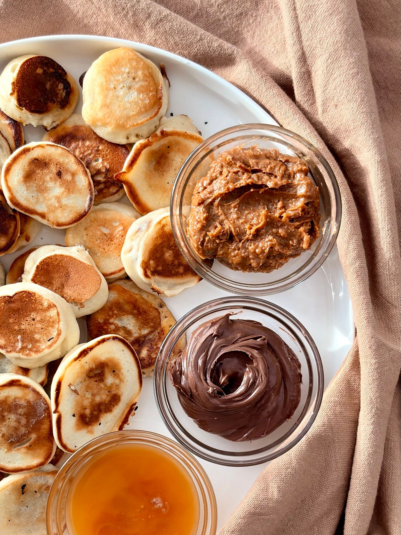 Overhead shot of mini pancakes on a white plate with three small glass bowls of toppings: honey, chocolate spread, and dulce de leche.