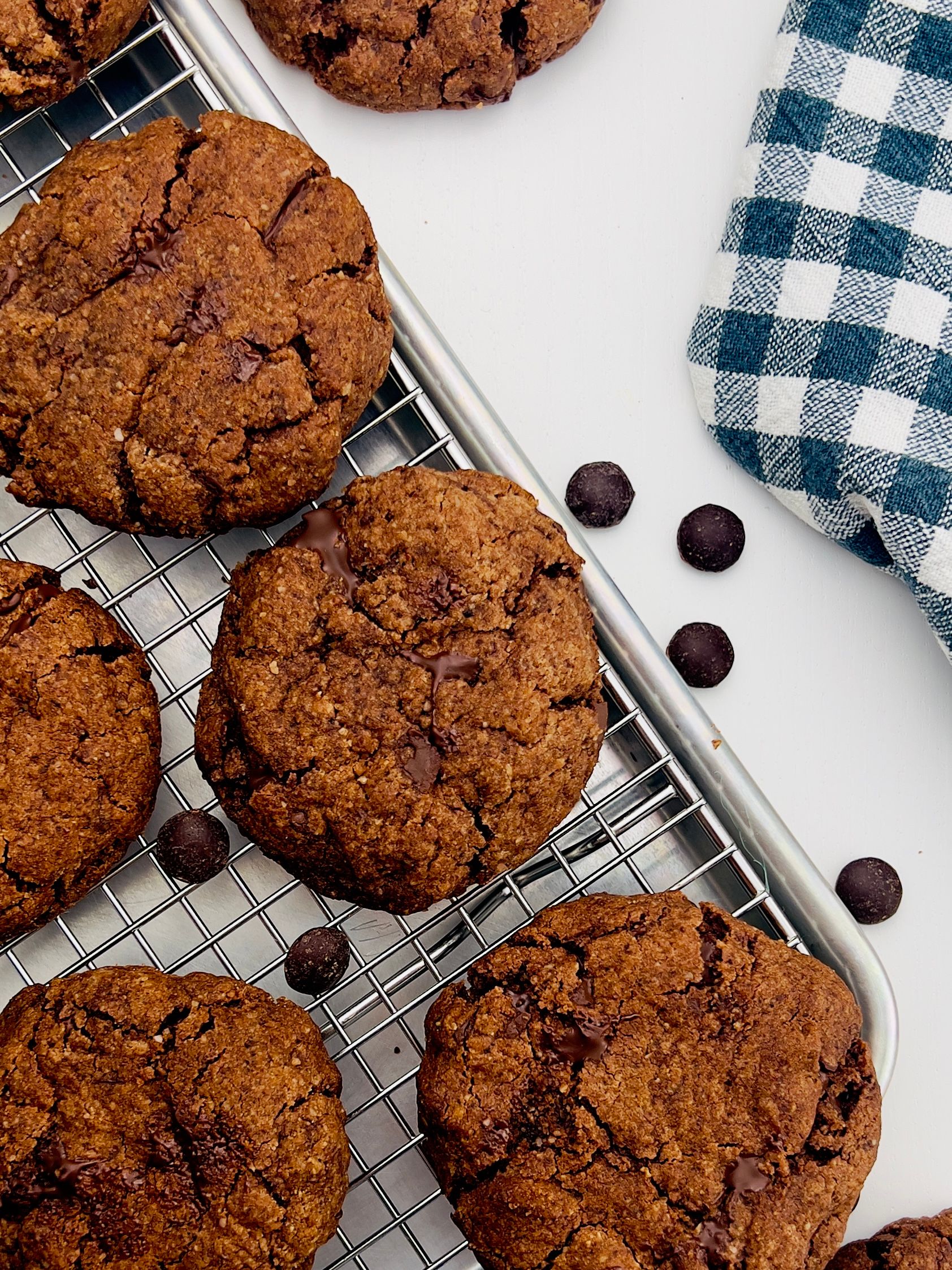 Close-up of five chocolate chip cookies, slightly cracked on top, cooling on a metal rack, with a blue and white checkered towel.