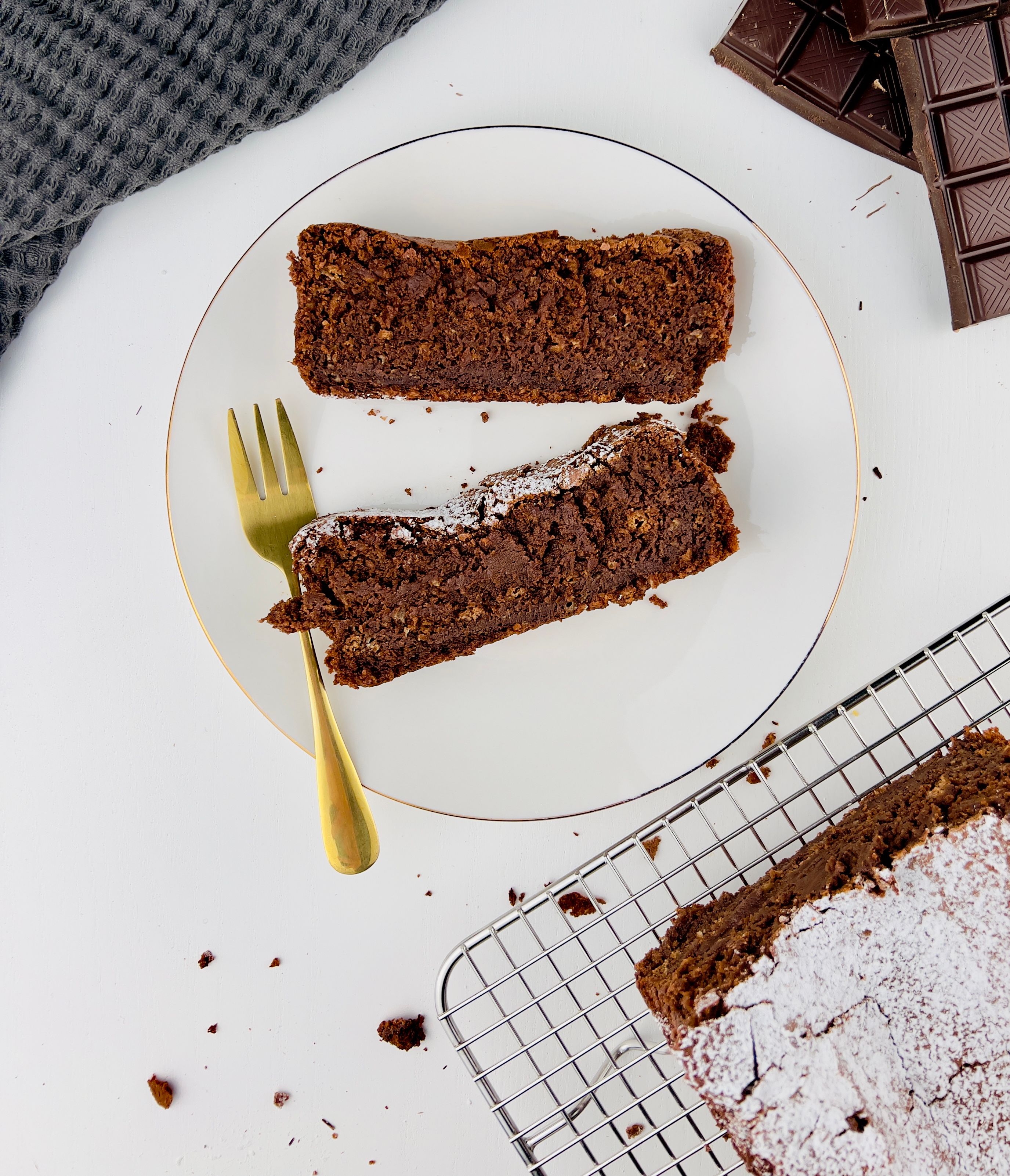 Two slices of chocolate bread cake with powdered sugar on a white plate with a gold fork, next to a cooling rack with more cake and dark chocolate bars, all on a white surface.