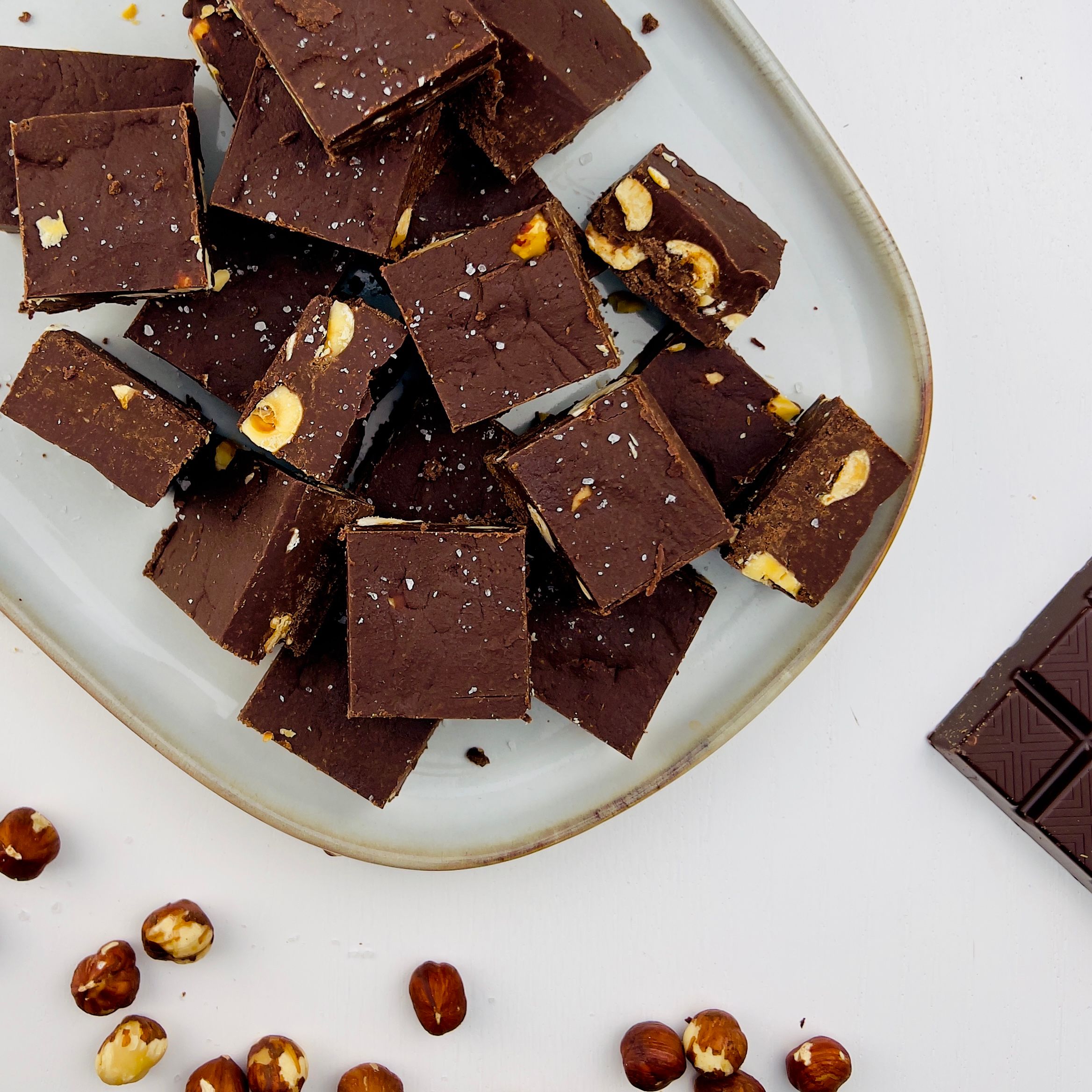 Overhead shot of chocolate fudge squares with hazelnuts on a plate. Hazelnuts and a chocolate bar are also on the white table.