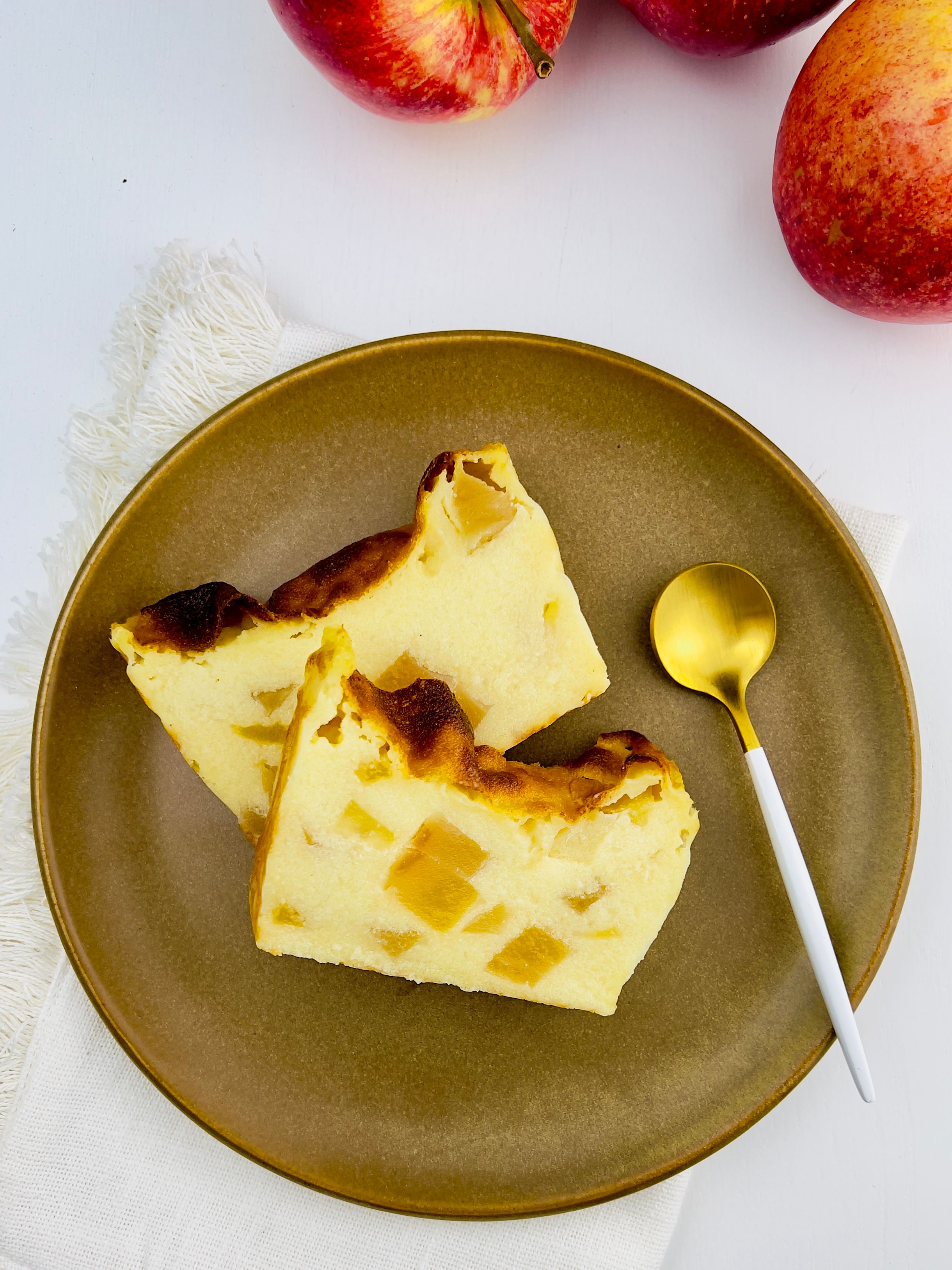 Overhead view of two slices of a custard-like semolina cake studded with apple chunks, on an olive-green plate with a gold spoon, red apples in the background.