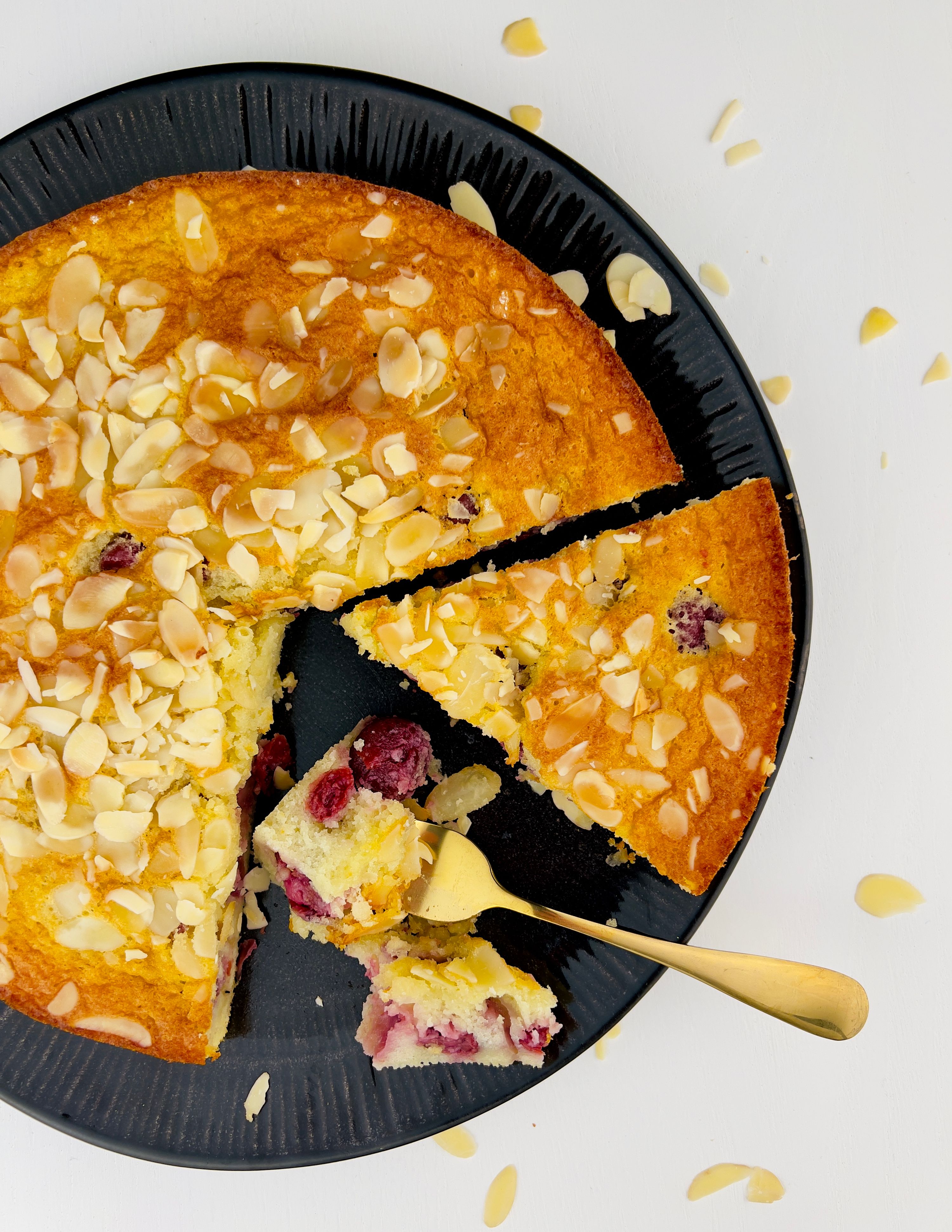 Close up, top-down view of a round yellow cake with a slice removed, revealing cranberries, topped with almond slivers