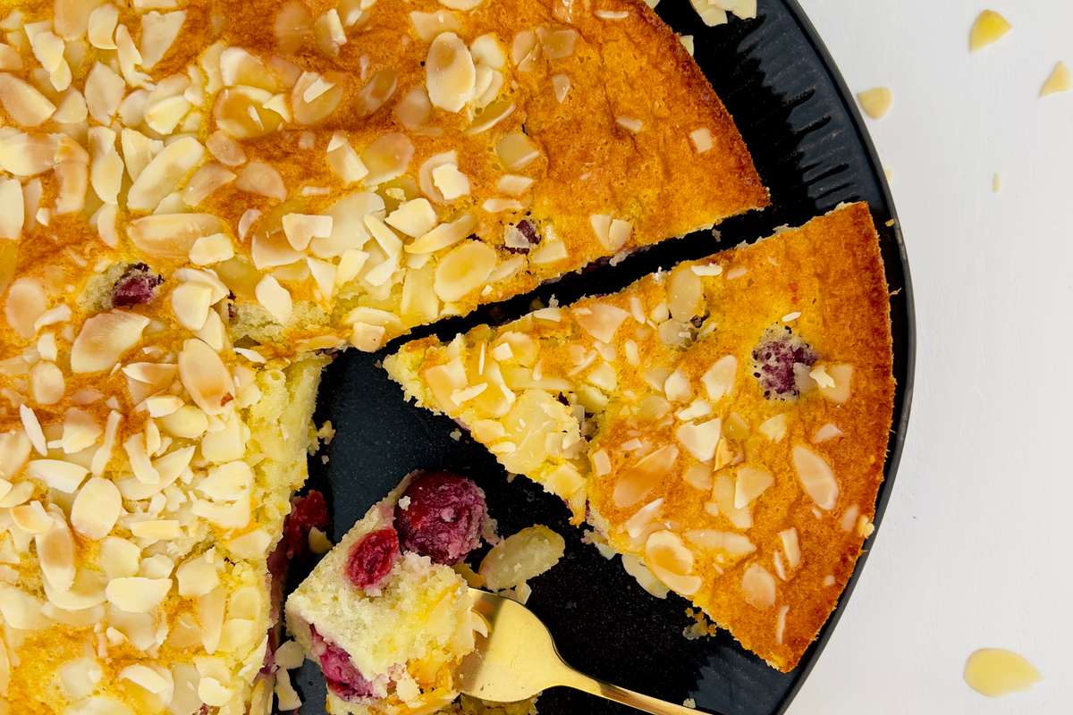 Close up, top-down view of a round yellow cake with a slice removed, revealing cranberries, topped with almond slivers