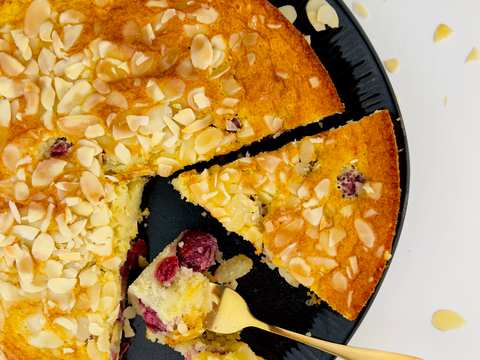 Close up, top-down view of a round yellow cake with a slice removed, revealing cranberries, topped with almond slivers