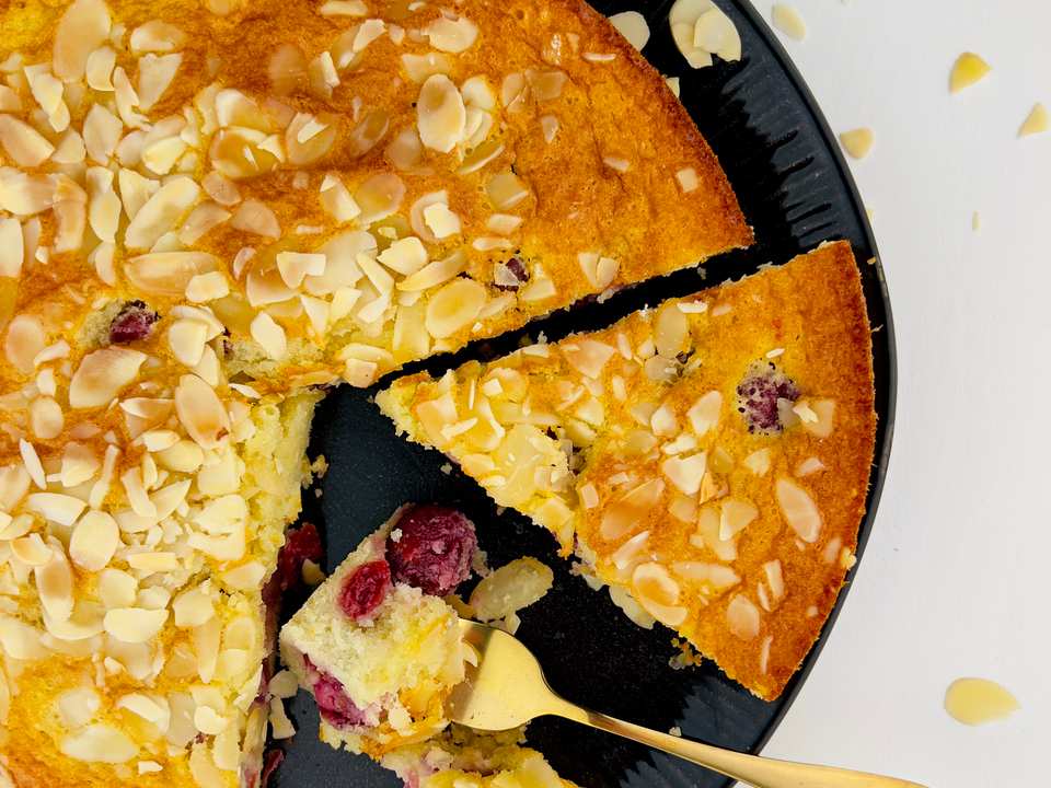 Vue rapprochée, de haut en bas, d'un gâteau rond et jaune avec une tranche retirée, révélant des canneberges, garni d'amandes effilées