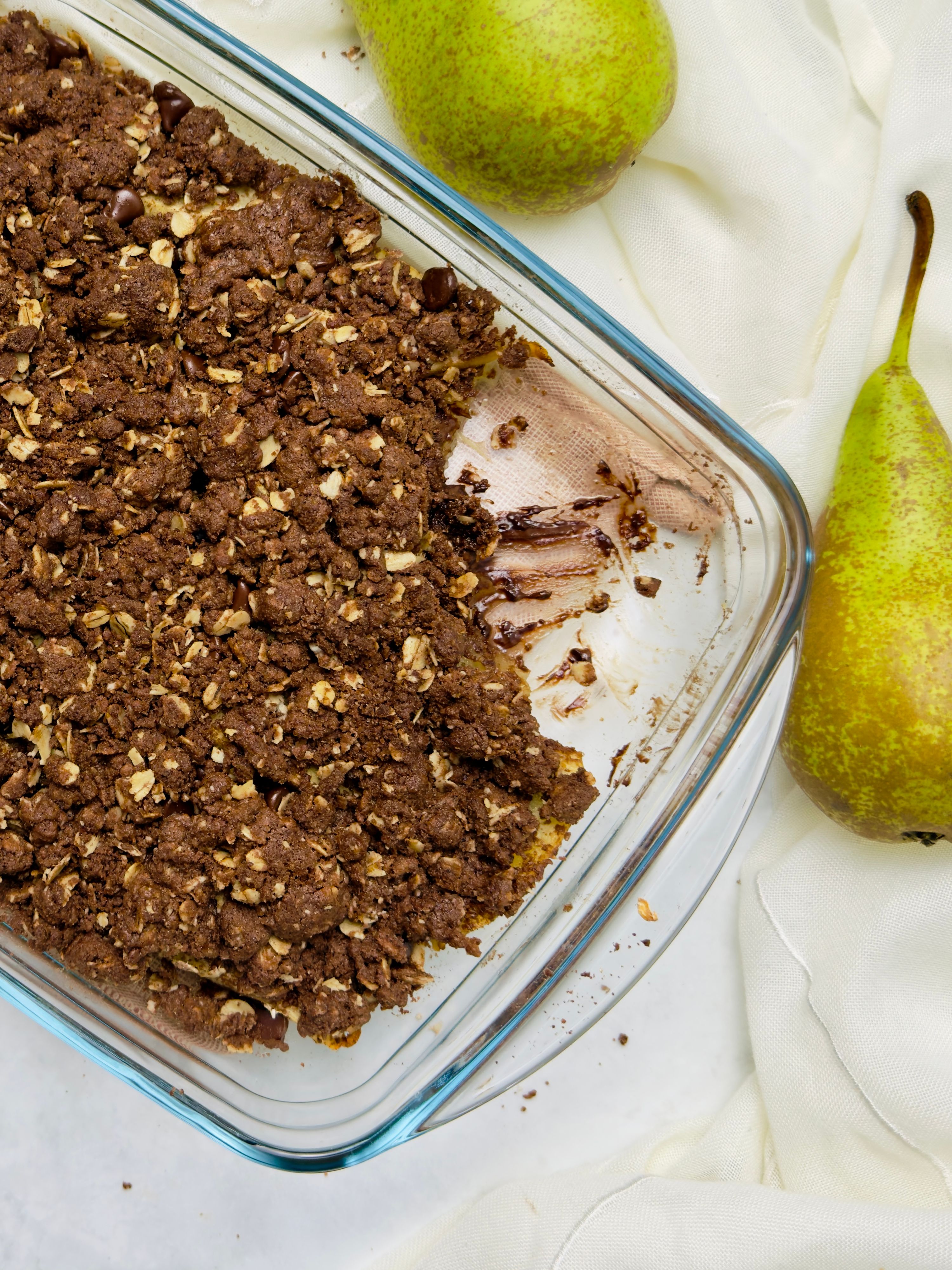 Close-up of a partially eaten chocolate pear crumble in a glass dish, with two pears and a white towel on the side.