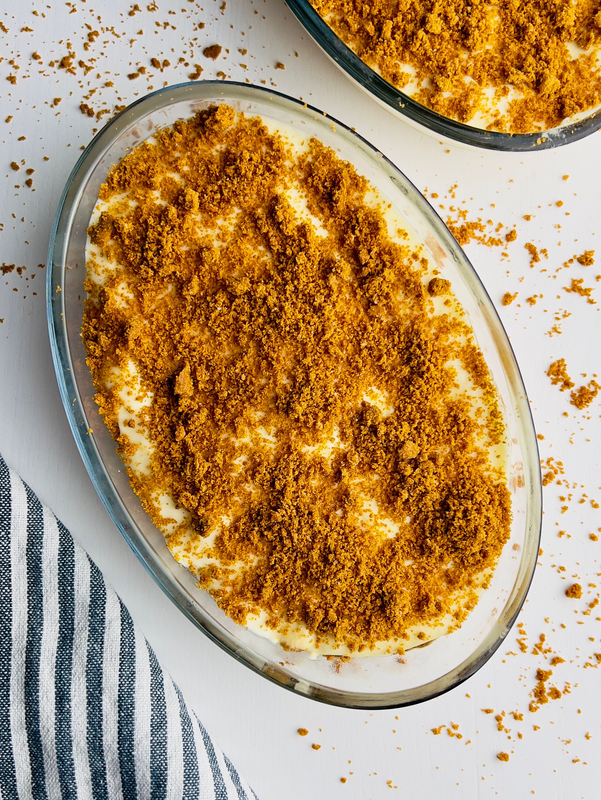 Two glass oval dishes, one in the foreground, filled with creamy white dessert and topped generously with light brown crumble. A striped napkin is to the lower left, and crumbs are scattered across the white surface.