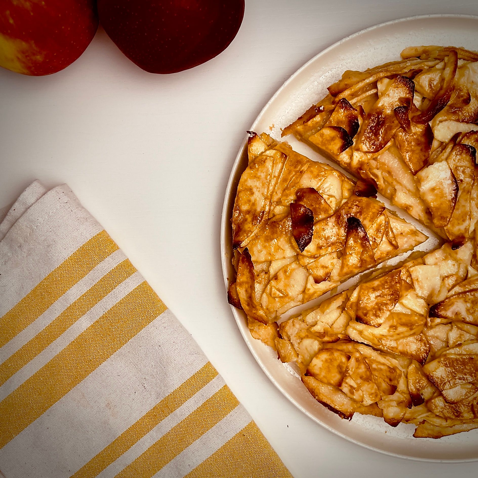 Rustic apple tart with several slices cut, showing golden-brown caramelized apples on a white platter, with two apples and a yellow striped linen next to it.