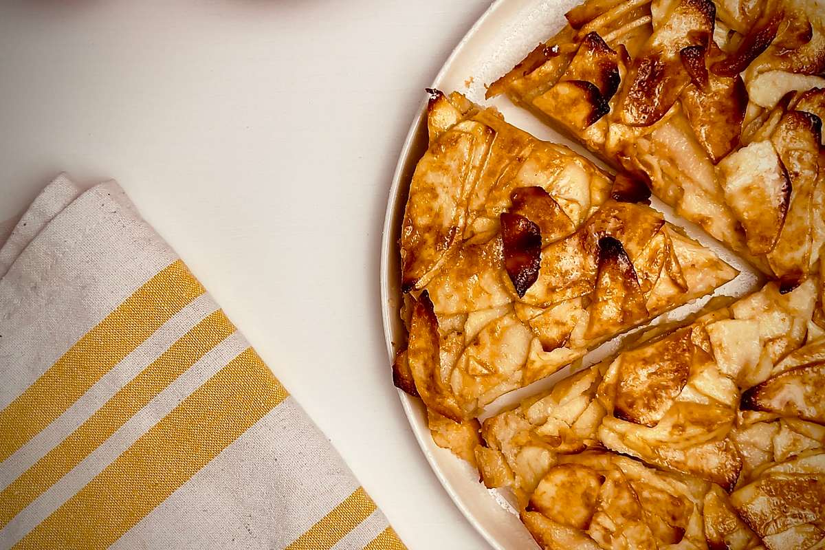 Tarte rustique aux pommes avec plusieurs tranches coupées, montrant des pommes caramélisées dorées sur un plat blanc, avec deux pommes et un linge à rayures jaunes à côté.