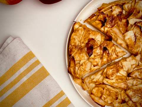Rustic apple tart with several slices cut, showing golden-brown caramelized apples on a white platter, with two apples and a yellow striped linen next to it.