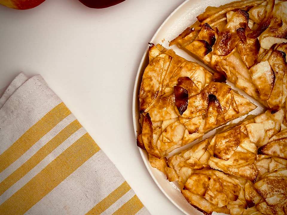 Rustic apple tart with several slices cut, showing golden-brown caramelized apples on a white platter, with two apples and a yellow striped linen next to it.