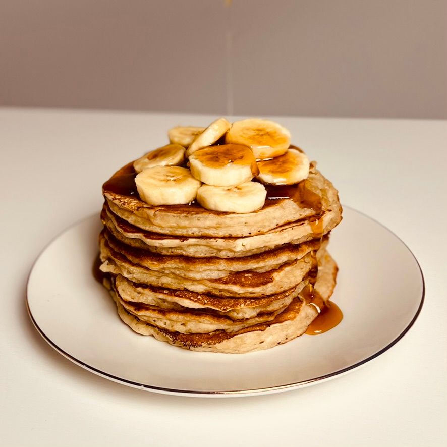 Stacked banana pancakes on a white plate. The pancakes are topped with sliced bananas and a drizzle of syrup.
